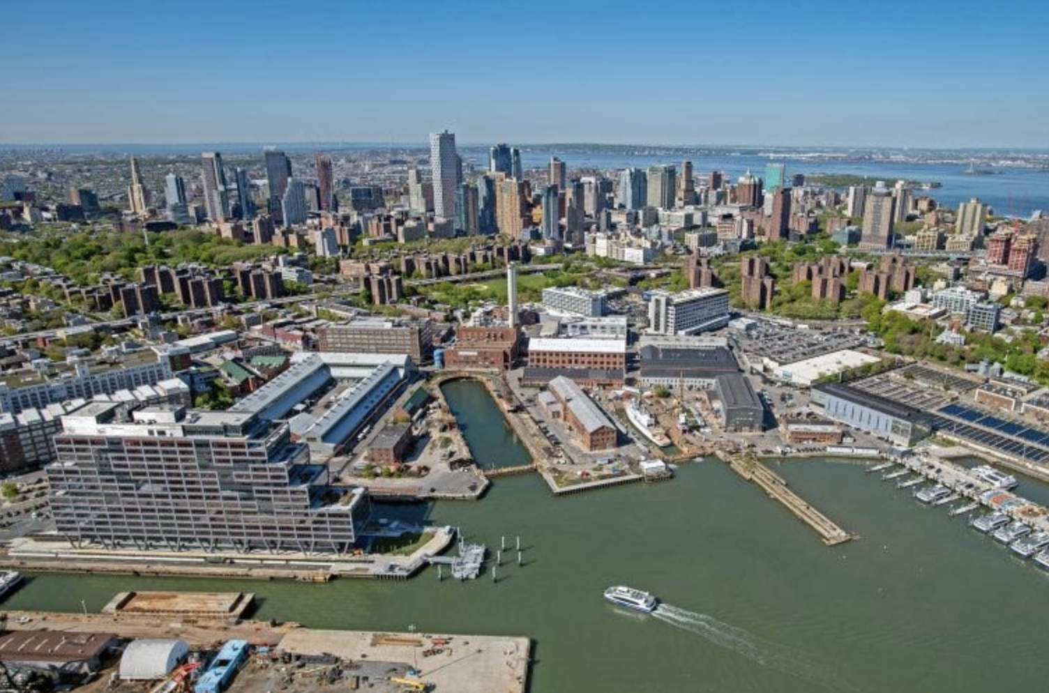 Aerial view of the Brooklyn Navy Yard campus along the East River with the Brooklyn skyline behind